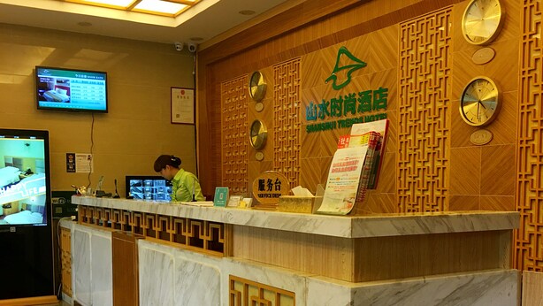 Een houten receptiebalie van een hotel, met meerdere klokken en een scherm dat kamertarieven toont.