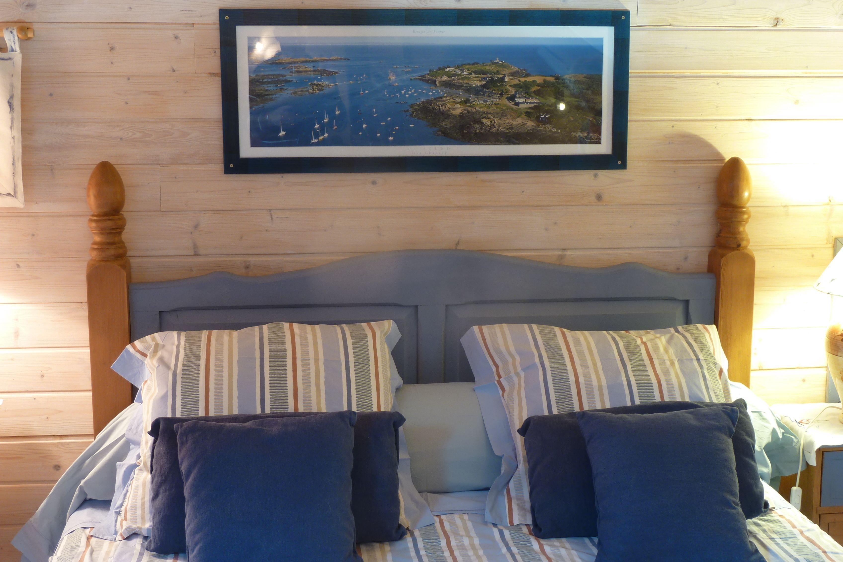 Un lit bien fait, avec une literie à rayures et deux oreillers, une photo encadrée représentant un paysage côtier au-dessus, et une tête de lit et un pied de lit en bois.