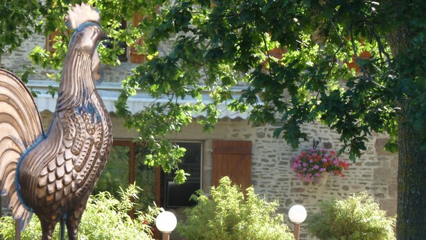 Una estatua de gallo en un jardín con plantas en macetas y un edificio de piedra al fondo.