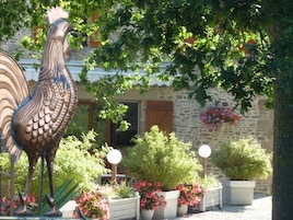Una estatua de gallo en un jardín con plantas en macetas y un edificio de piedra al fondo.