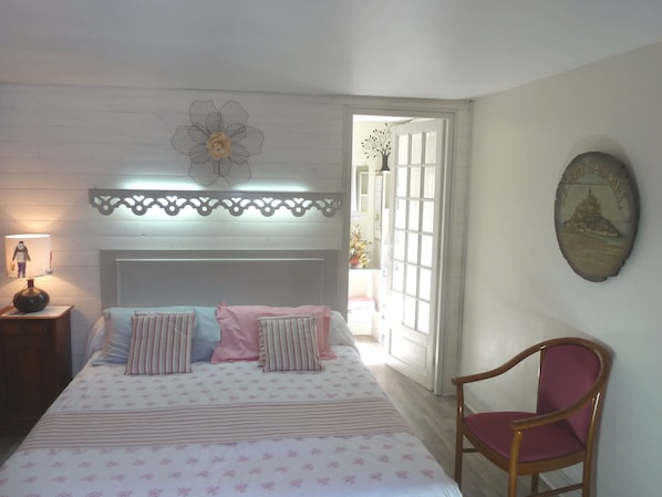 Un dormitorio con cama, una silla, una mesita de noche, lámpara, un adorno de pared y ventana.