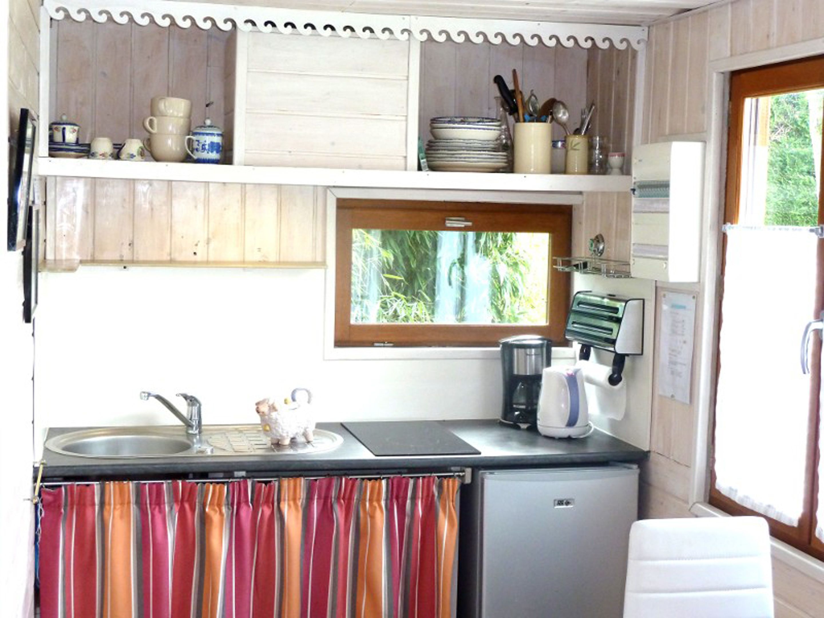 A compact kitchen with a sink, a coffee maker, and a small refrigerator. There is a window above the sink providing natural light, and a shelf with dishes and utensils.