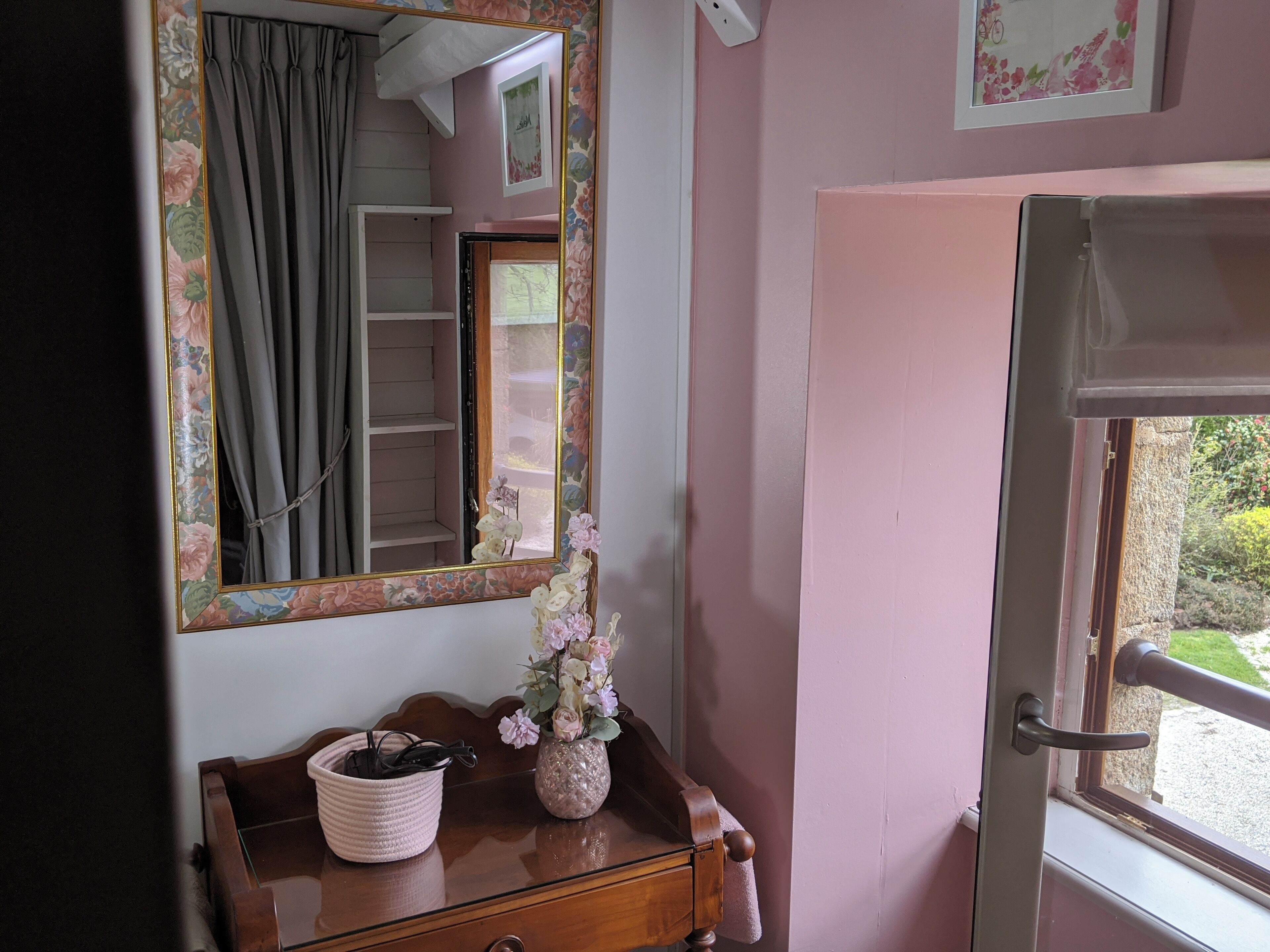 A room with a wooden dresser, a mirror with a floral frame, a vase with flowers, and a door leading outside.