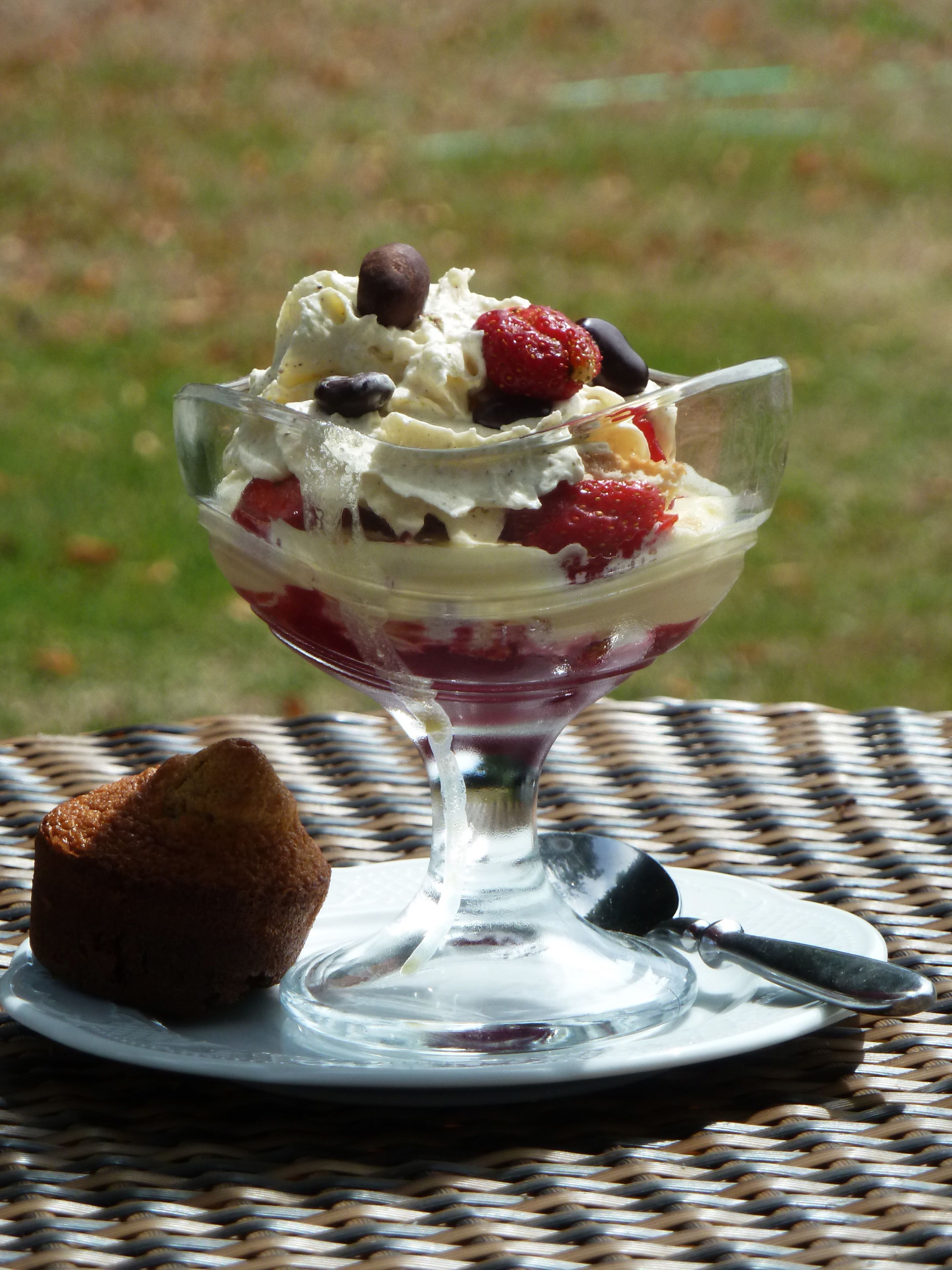 Un dessert servi dans un bol en verre, garni de crème fouettée et de fruits rouges, accompagné d’un muffin posé sur une nappe à motifs tissés.