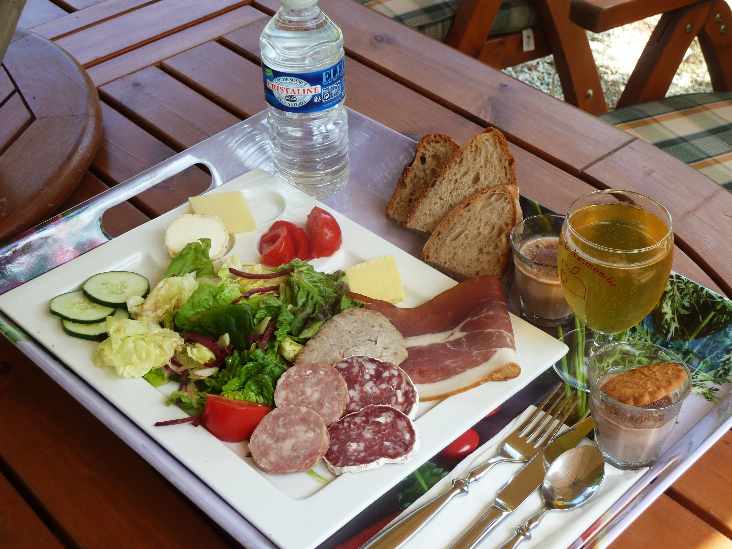 A meal tray with a variety of foods including bread, cheese, meats, and vegetables, along with a bottle of water and a glass of juice.