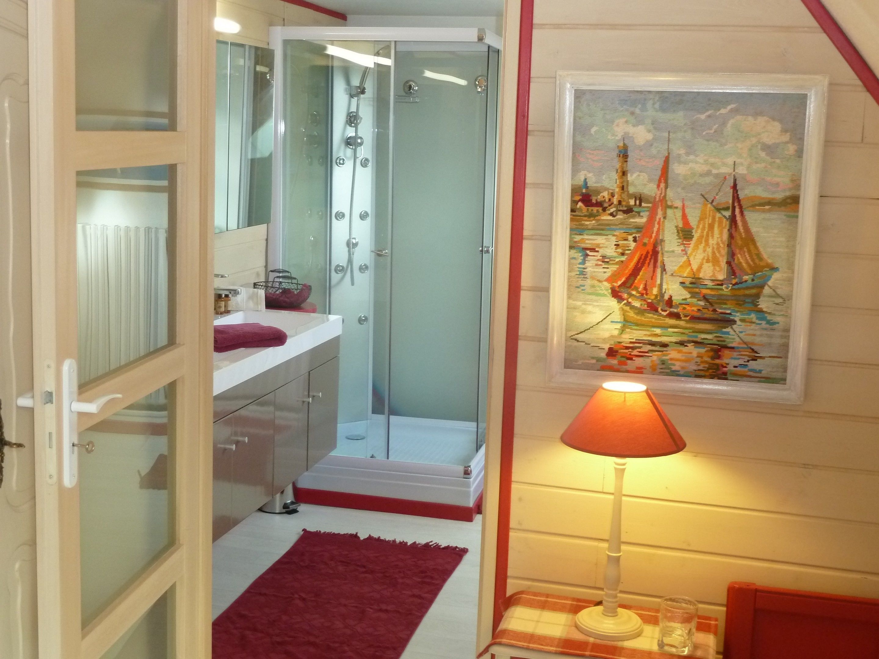 Une salle de bain avec une douche à parois de verre, un lavabo avec un miroir, un tapis rouge et un tableau représentant des voiliers accroché au mur.