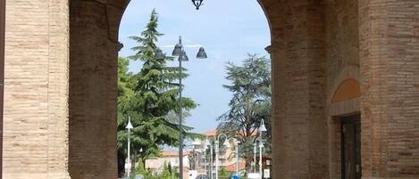 An arched stone gateway with a hanging lantern, leading to a street with directional signs and trees.