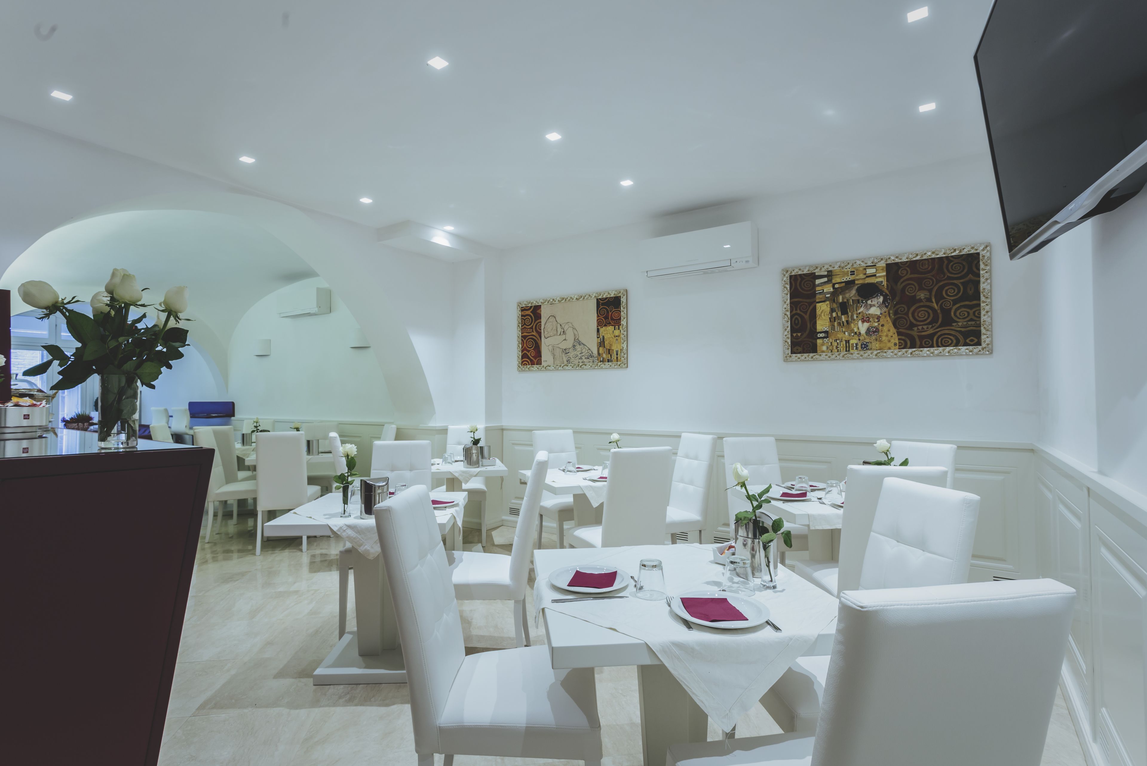 Zona pranzo con tavoli e sedie bianche, un vaso con fiori e due quadri alle pareti.
