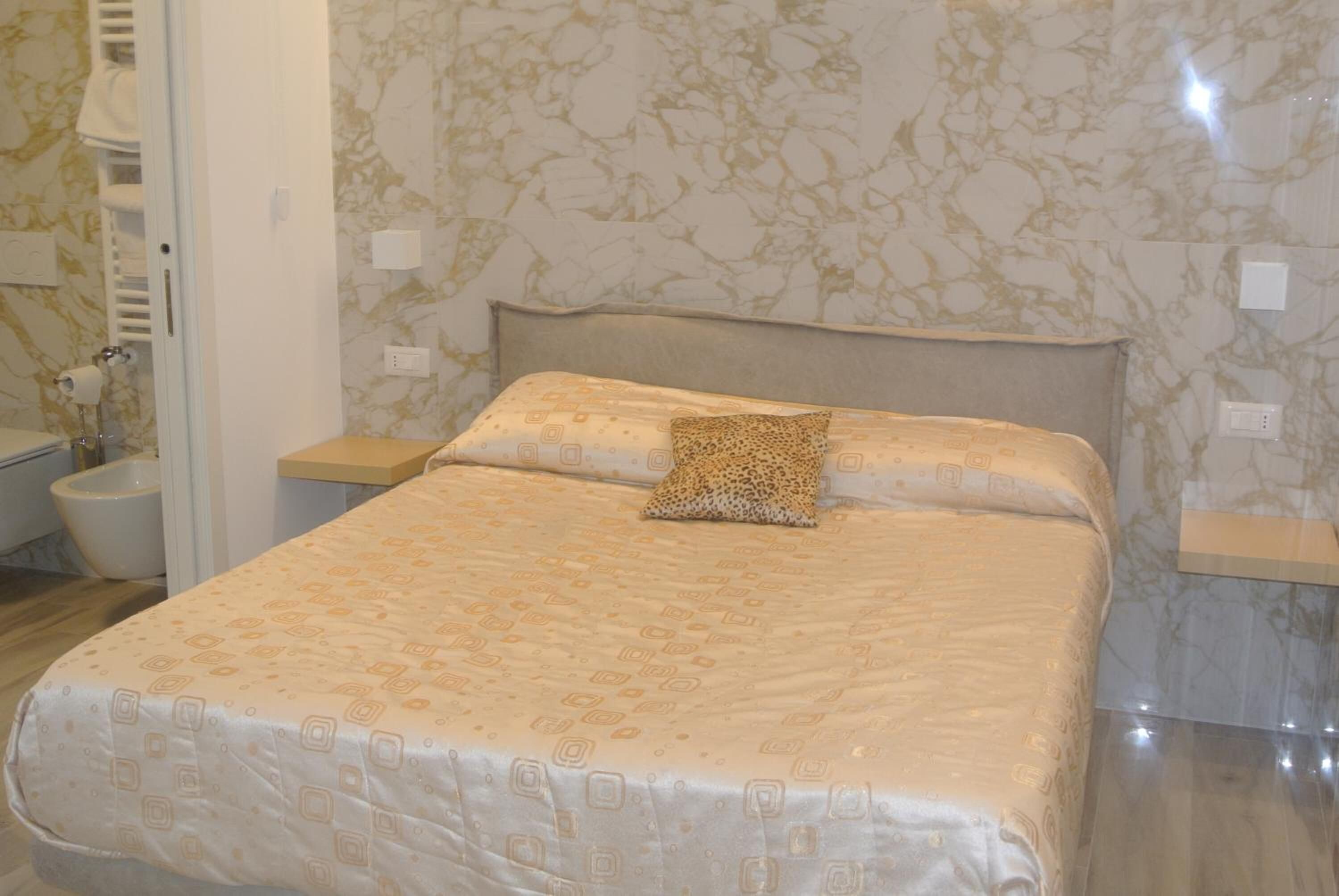 Ein ordentlich bezogenes Bett mit einem gemusterten Bettbezug und einem leopardenprint Kissen, eine weiße Wand mit marmorartigem Muster und ein kleiner Nachttisch mit einer Lampe.