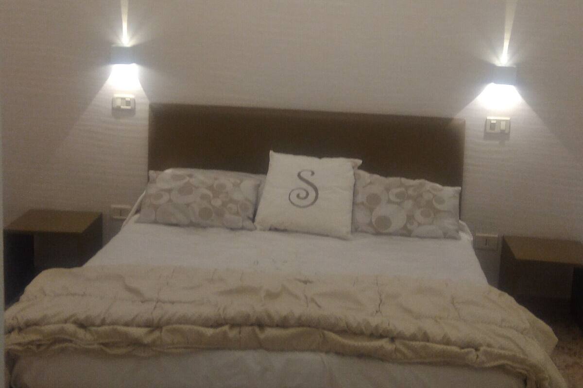 A neatly made bed with a pillow marked with an 'S', two bedside lamps, and a carpeted floor.