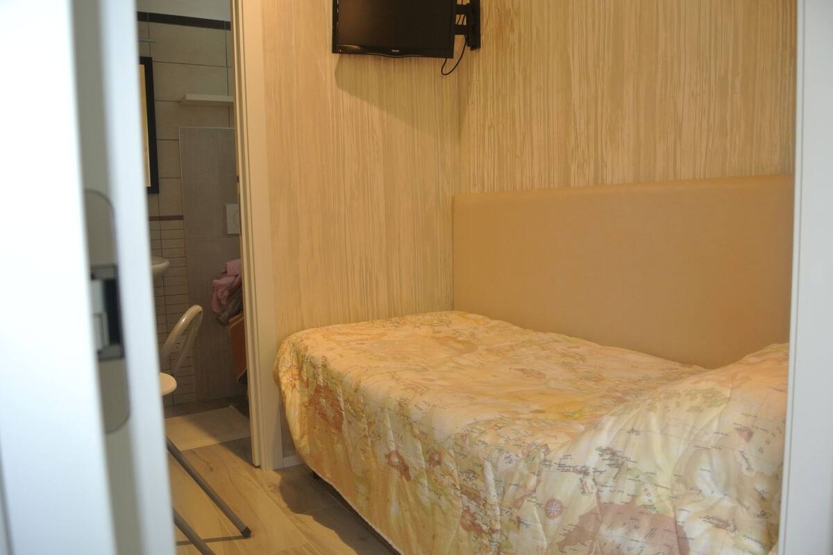 A small, single-bed room with a wooden headboard, a visible bathroom with a toilet and a shower, and a wall-mounted TV.