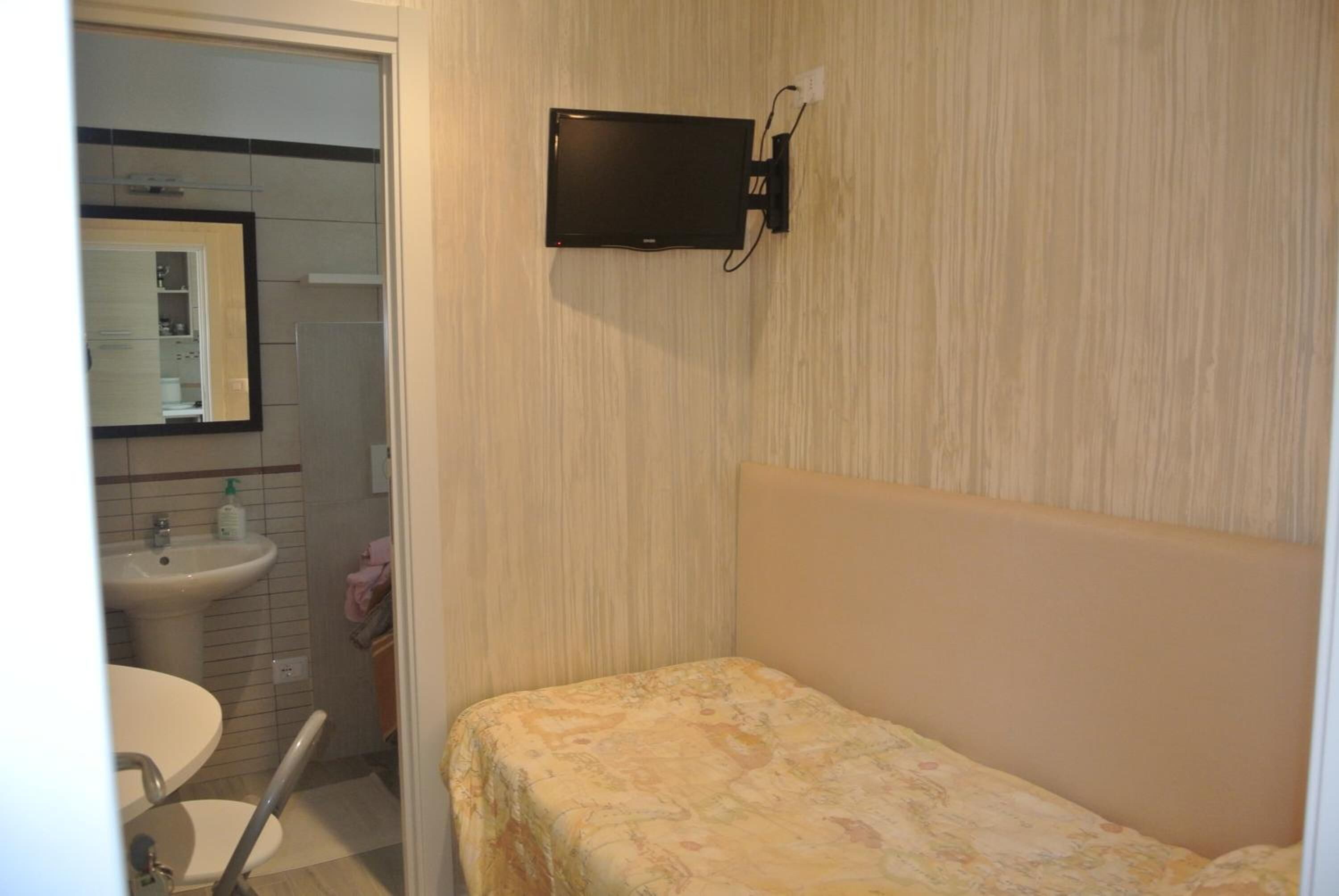 A small, single-bed room with a wooden headboard, a bedside table, and a mounted flat-screen TV.
