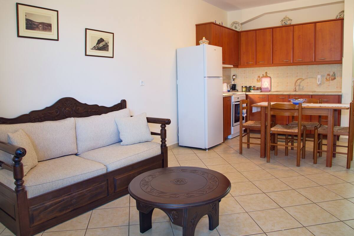 Uma sala de estar com sofá, mesa de centro e área de cozinha com armários de madeira e geladeira.
