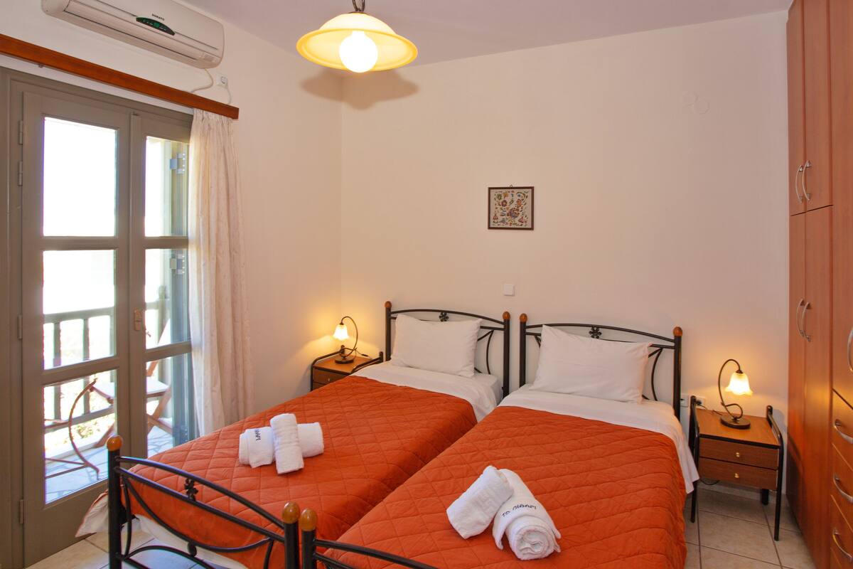Quarto de hotel com duas camas, um guarda-roupa de madeira, uma escrivaninha com cadeira e uma janela com cortinas.
