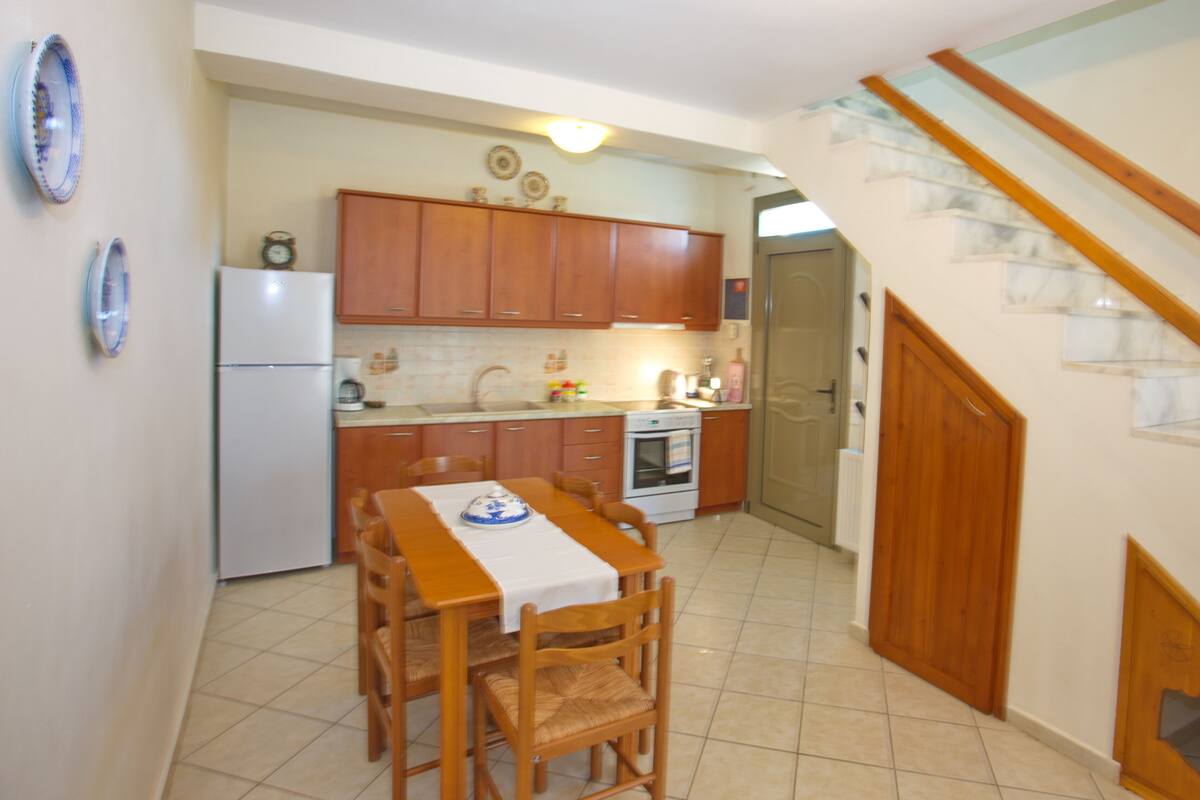 Uma cozinha com armários de madeira, uma mesa de jantar com cadeiras, uma geladeira e uma escada que leva a um andar superior.