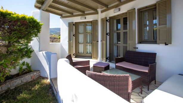 Un espacio cubierto al aire libre con mobiliario de mimbre y una mesa de vidrio, junto a un edificio con contraventanas de madera y una jardinera de piedra.