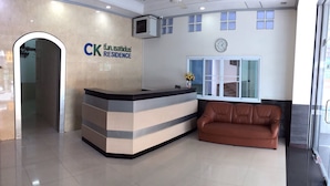 A reception area with a reception desk, a brown leather couch, a clock on the wall, and a window with blinds. - CK Residence (Hat Yai)