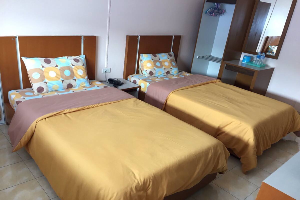 A hotel room with two beds, each with a patterned pillow and a brown bedspread. There is a bedside table with a phone and a small mirror.