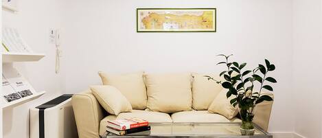 Una sala de estar con un sofá color beige, una mesa de centro de vidrio, una planta y un cuadro en la pared.