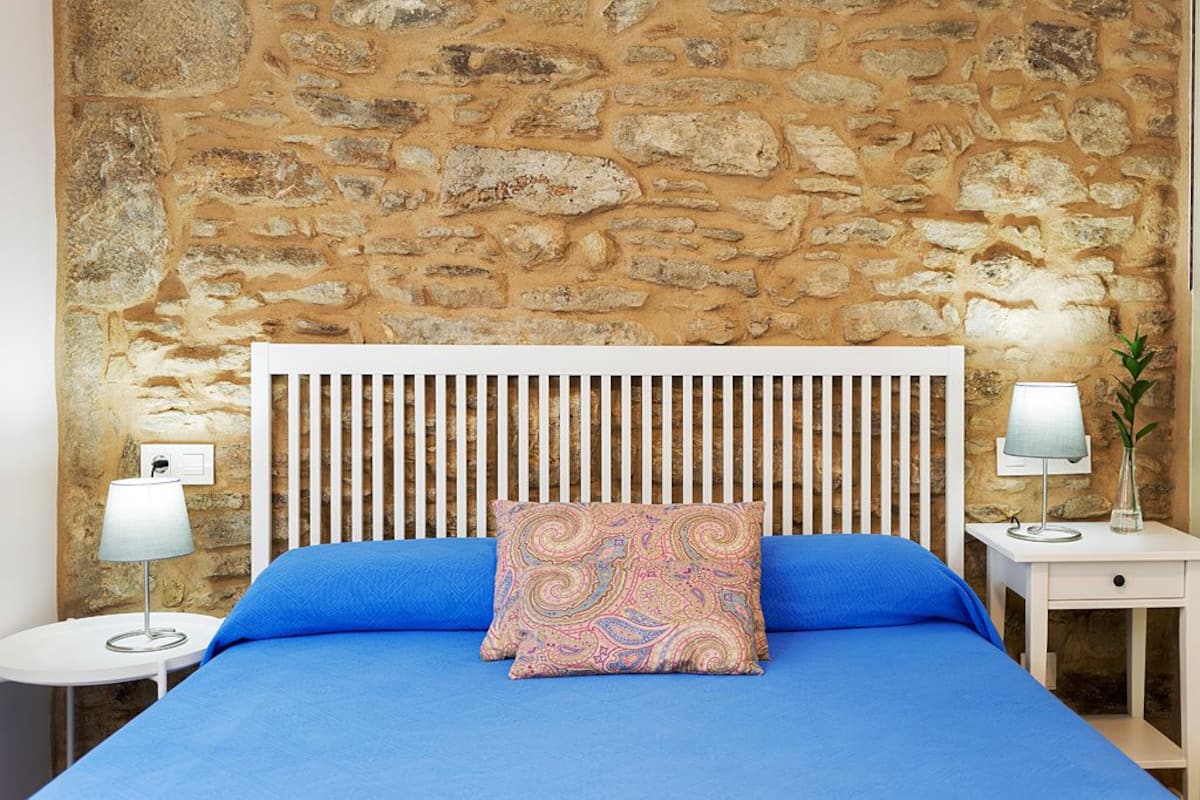 A bed with a white headboard, blue bedding, and a patterned pillow against a stone wall. There are two bedside tables with lamps and a small drawer.