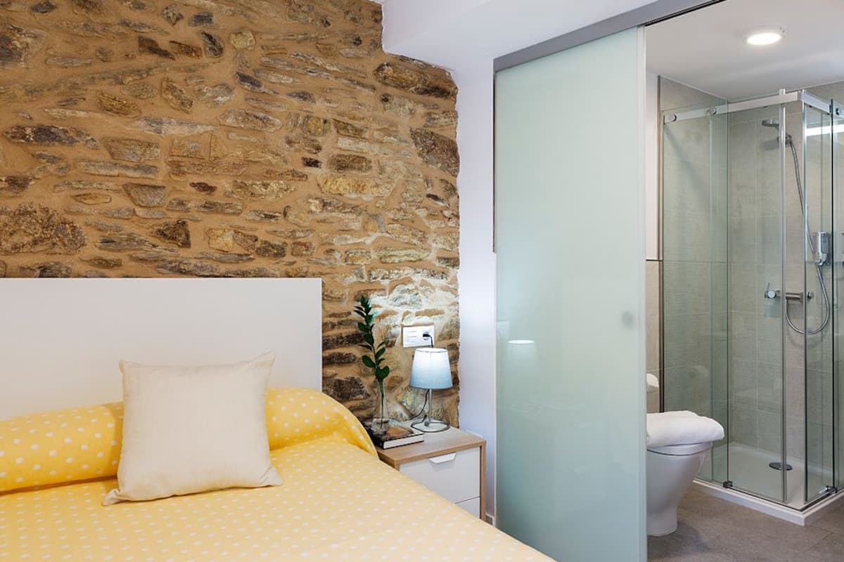 A bedroom with a stone wall, a bed with yellow bedding, a bedside table with a lamp, and a glass-enclosed shower area.