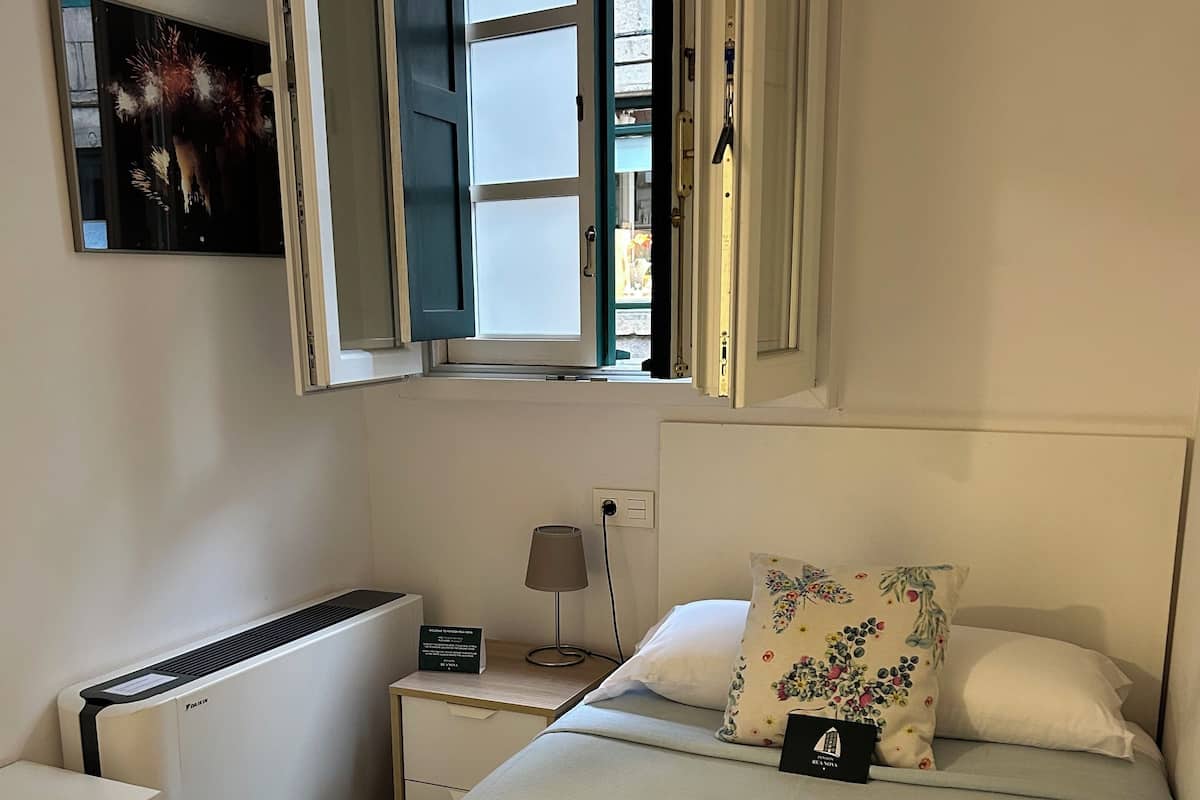 A hotel room with a bed, a desk, a radiator, a window, and a wall-mounted TV.