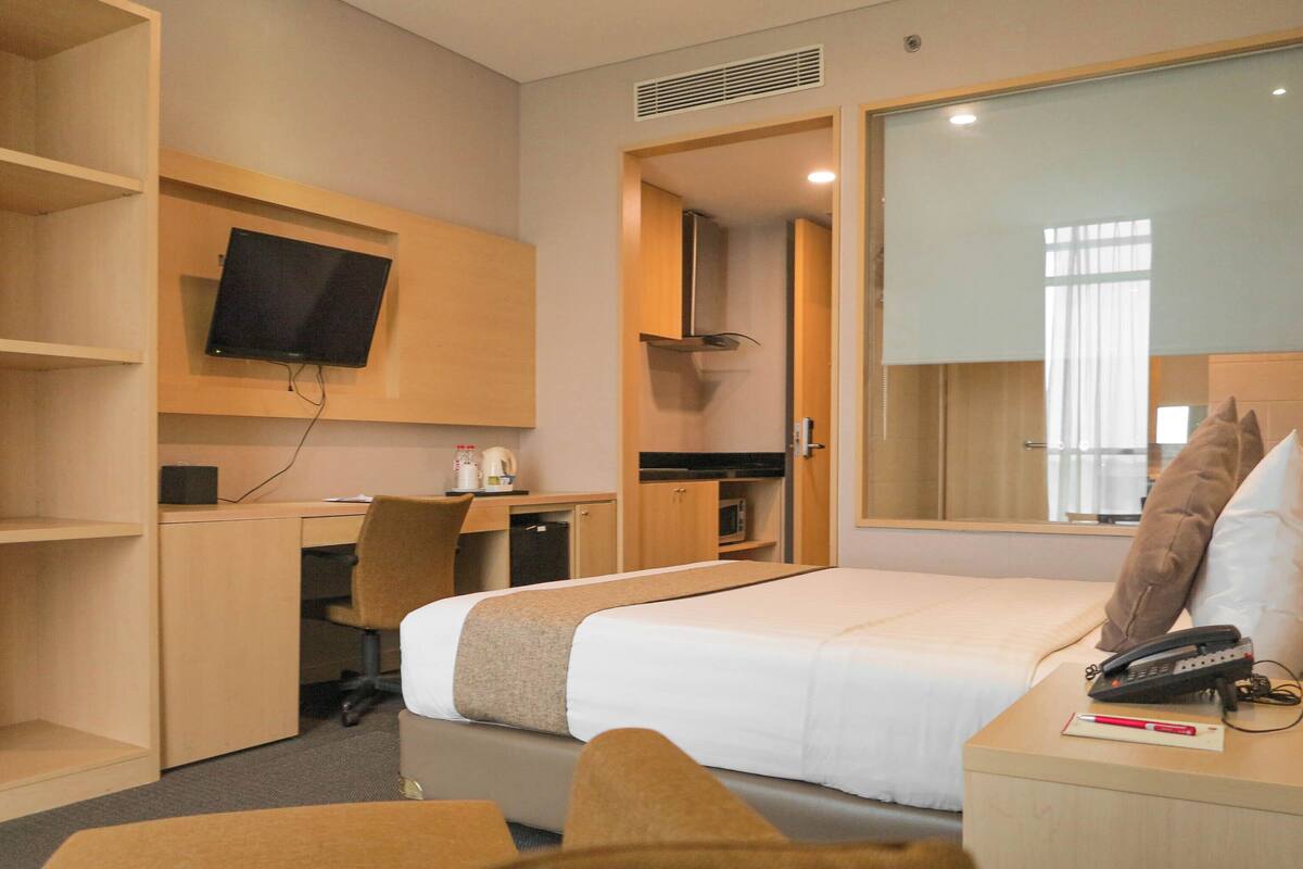 Habitación de hotel con cama, escritorio, silla, televisor, teléfono y ventana con cortinas.