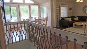 Ampla sala de estar com piso de madeira, sofá de cor escura, mesa de centro e mesa de jantar com cadeiras. Grandes janelas oferecem vista para o exterior.
