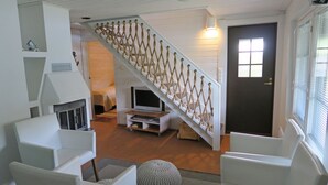 Uma sala de estar com móveis brancos, escada com corrimãos de corda, televisão sobre um suporte e uma porta com janela.