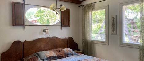 Une chambre à coucher avec un lit en bois, une table de chevet, une lampe fixée au mur, une fenêtre avec des rideaux et un miroir décoratif.