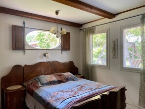 A bedroom with a wooden bed, a nightstand, a wall-mounted lamp, a window with curtains, and a decorative mirror. - Villa Prud (Kuopio)