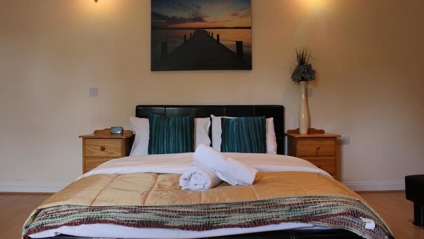 A neatly made bed with a striped blanket, two bedside tables with lamps, a painting of a sunset on the wall, and a tall plant on one table. - Remarc House Stansted (Bishop's Stortford)