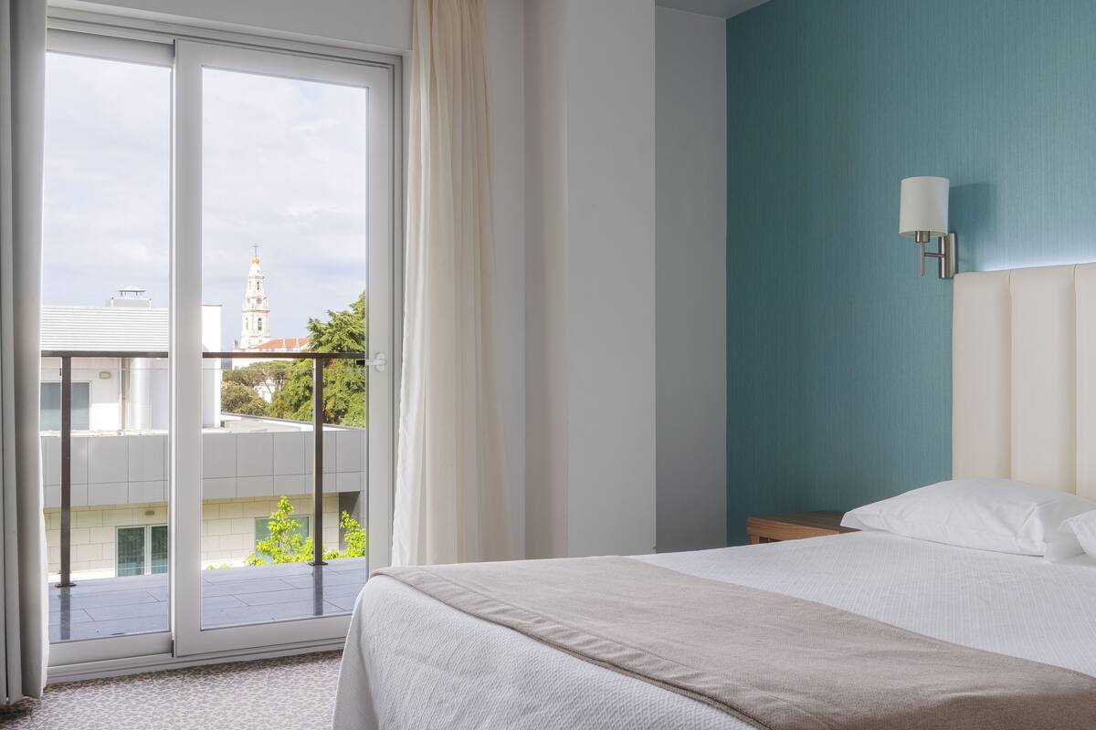 A hotel room with a bed, a nightstand, a wall-mounted lamp, and a view of a building through a large window.