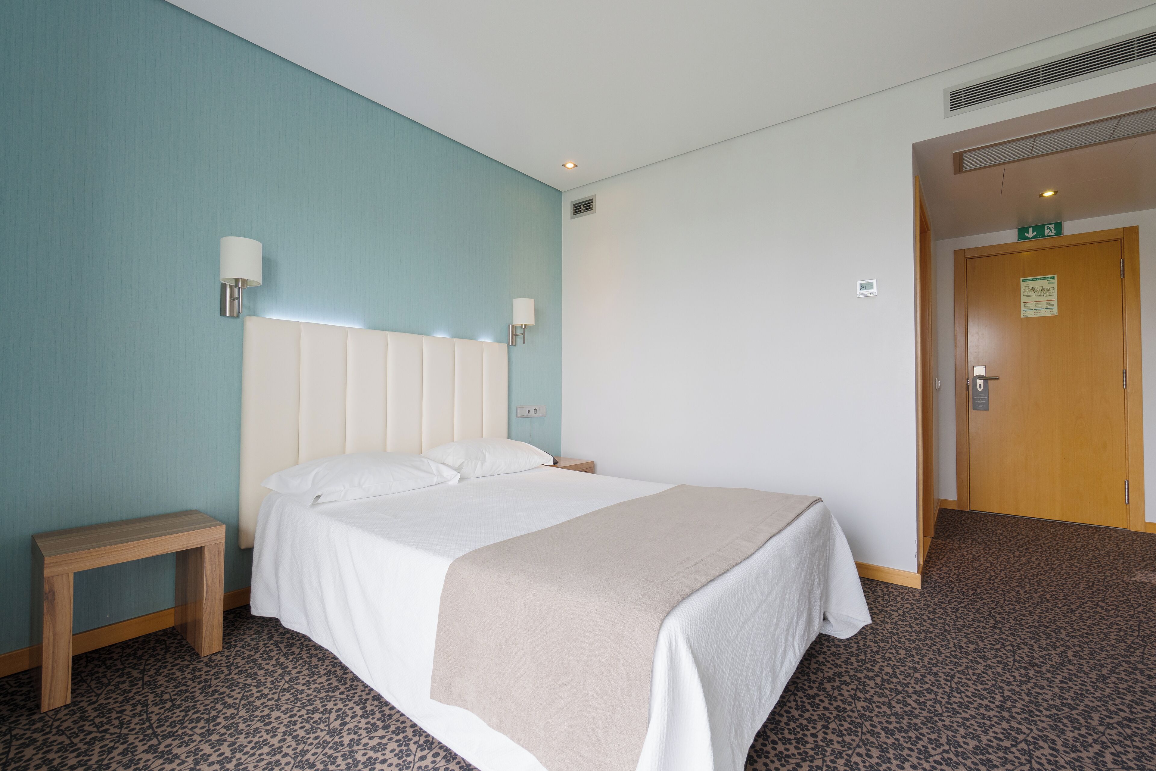 Habitación de hotel con cama, mesita de noche y lámpara de pared.