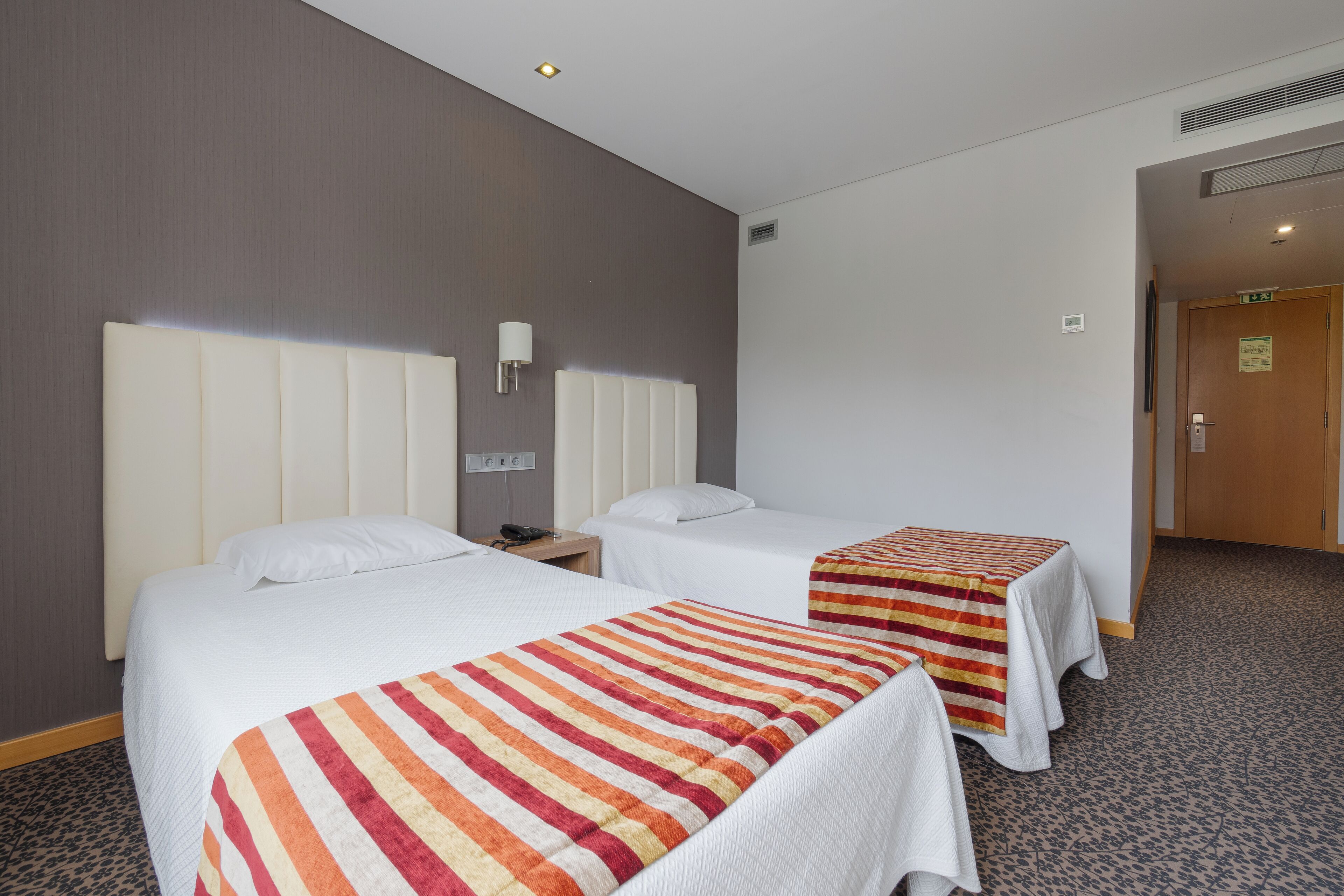 A hotel room with two beds, each with a striped blanket, a bedside table with a lamp, and a wall-mounted light.
