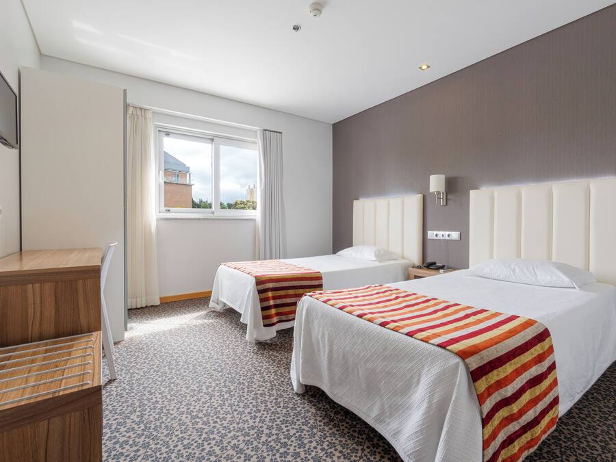 A hotel room with two beds, a desk, a TV, and a window with curtains.