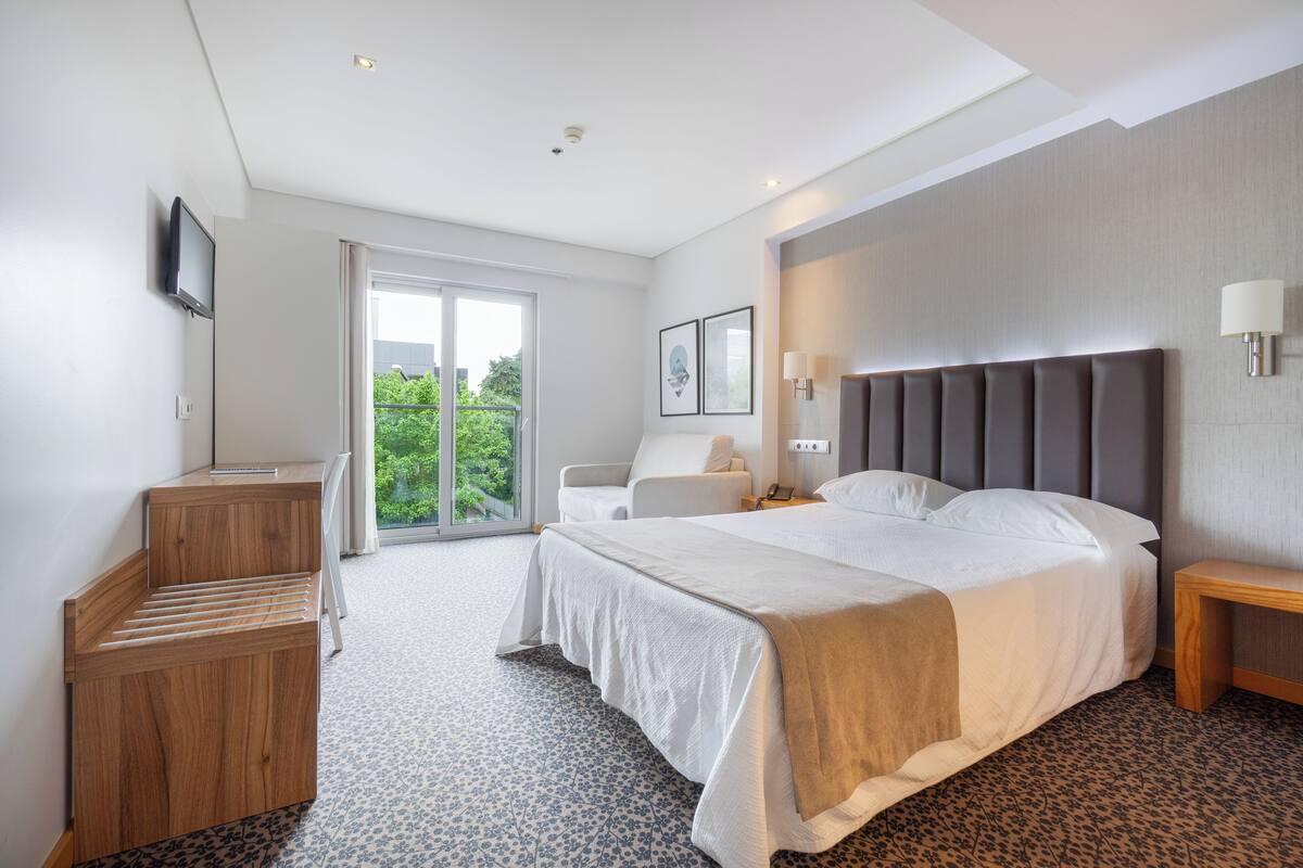 A modern hotel room with a large bed, a desk, a chair, a TV mounted on the wall, and a window with a view of greenery.