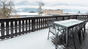 Een sneeuwbedekt terras met een tafel en stoelen, met uitzicht op een besneeerd landschap en gebouwen.