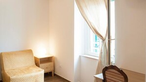 Una habitación pequeña y poco amueblada con una sola silla y una mesita, una ventana con cortinas y vistas a edificios.