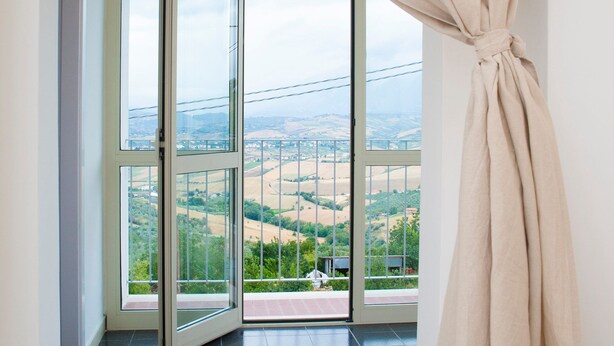 Una habitación con un gran ventanal corredera que da a un balcón con vistas a un valle y edificios lejanos.