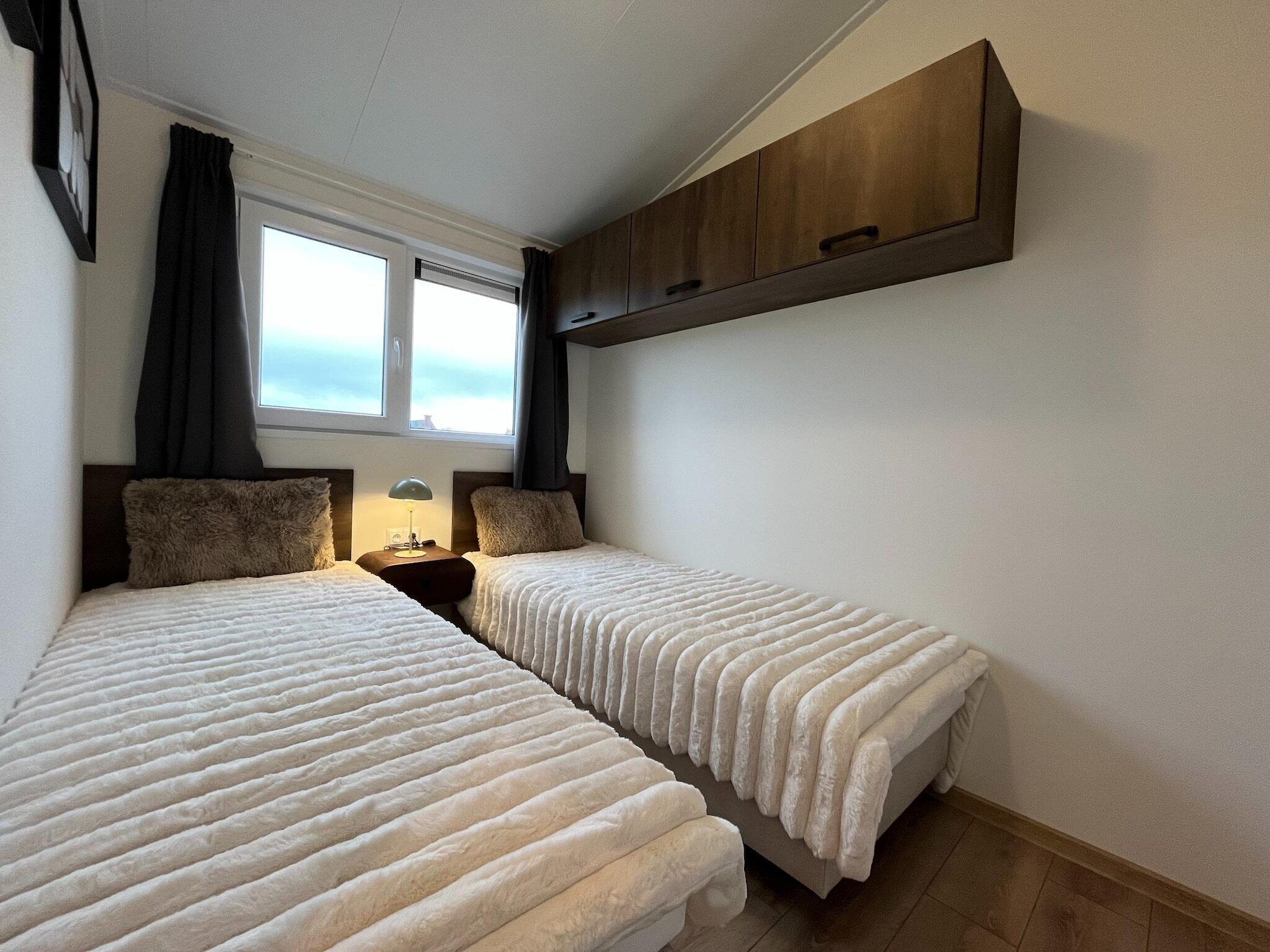 A bedroom with two beds, a nightstand, a wall-mounted cabinet, and a window with curtains.