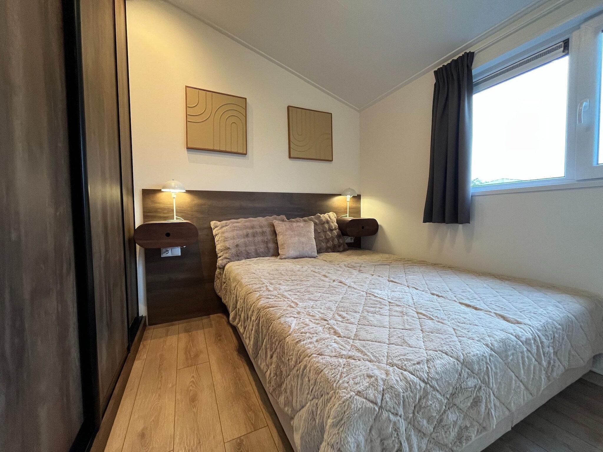 Quarto com cama, duas luminárias fixadas na parede, janela com cortinas e uma pequena mesa de cabeceira de madeira.