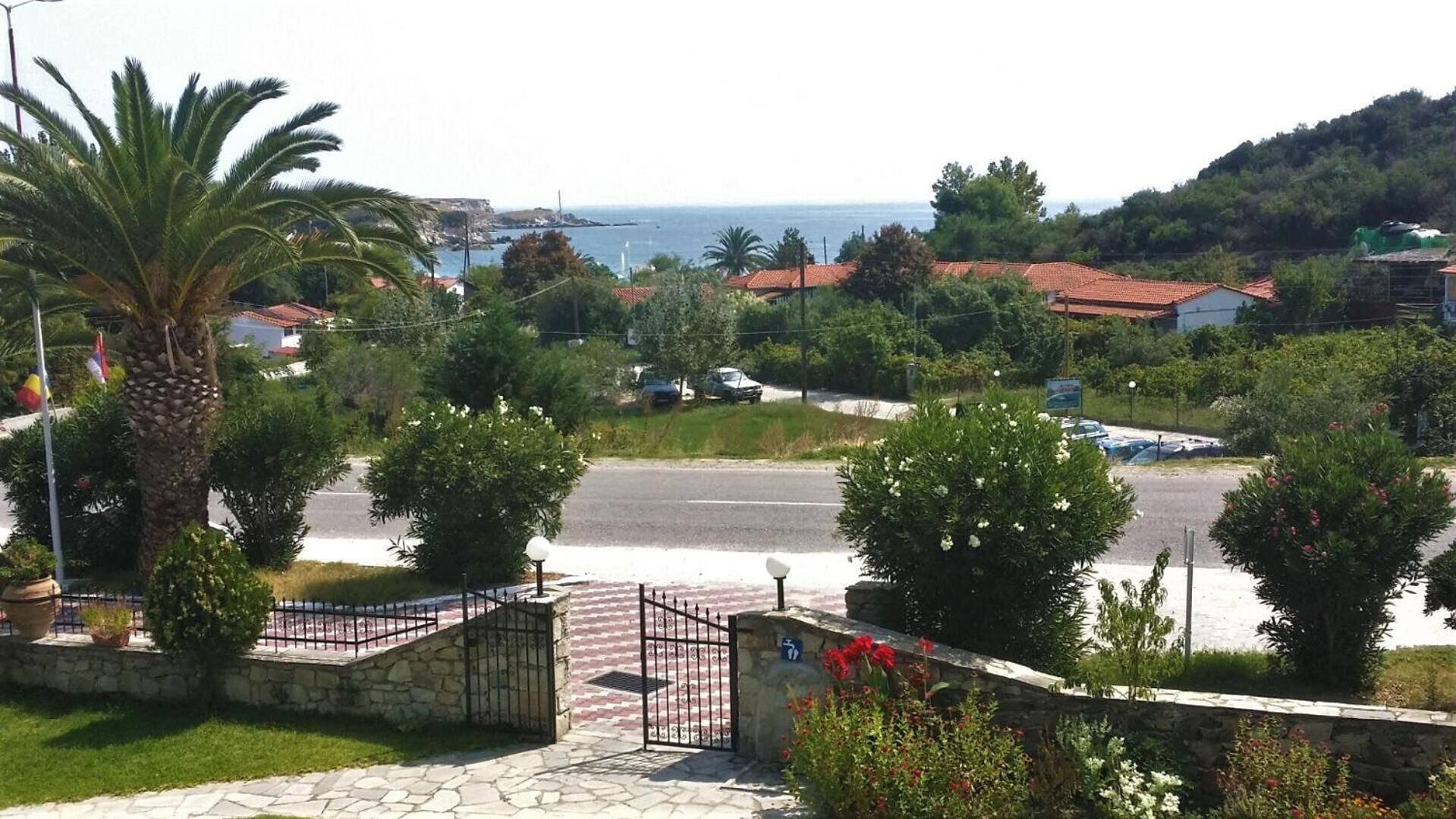 A coastal road with a stone entrance gate, palm trees, and a view of the sea and hills in the background.