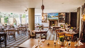 A restaurant with checkered flooring, wooden tables, and chairs. There are hanging light fixtures and a fireplace. - Crown Inn at Pooley Bridge (Penrith)
