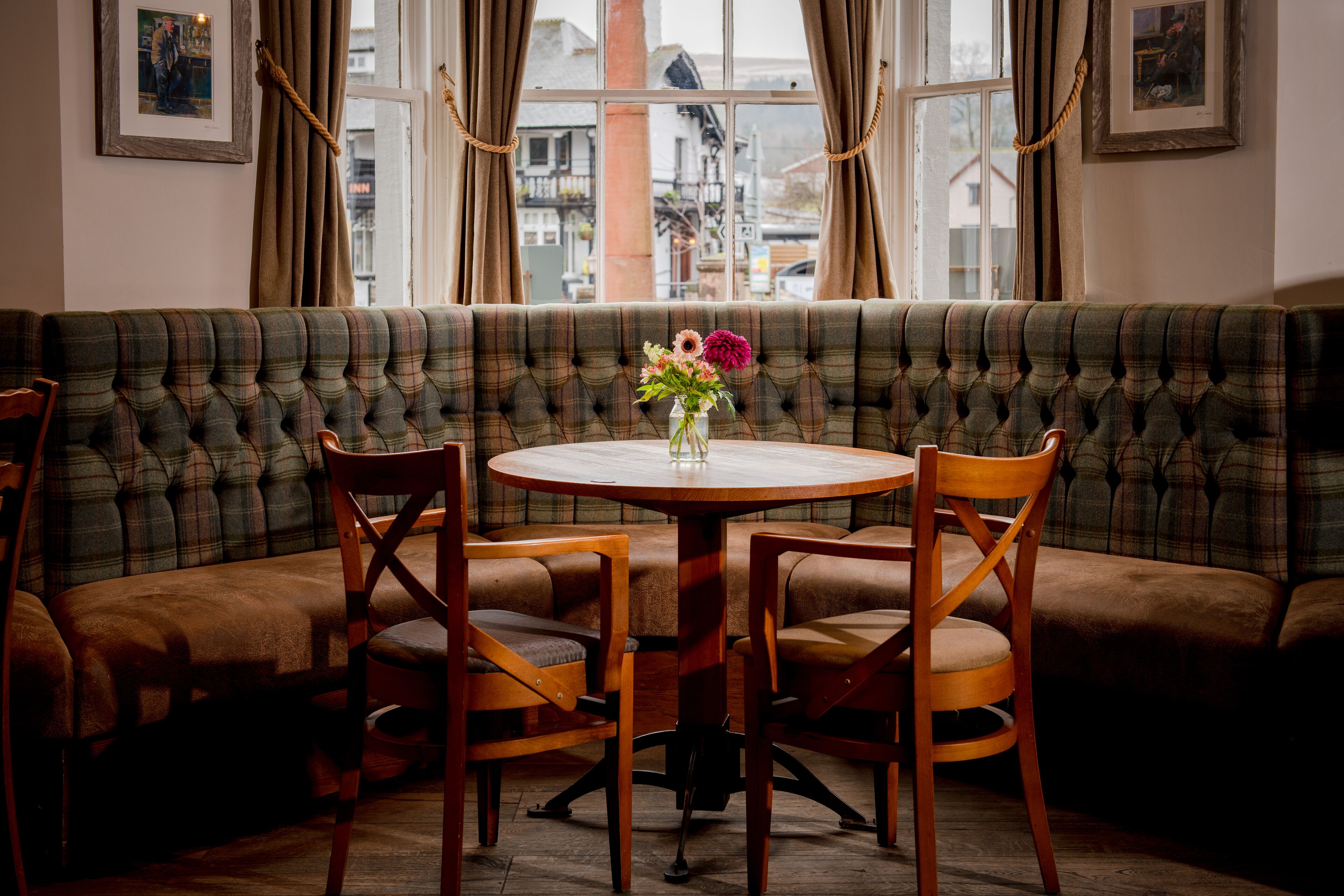 A cozy seating area with a round wooden table and four chairs, a vase with flowers, and a view of buildings through large windows.