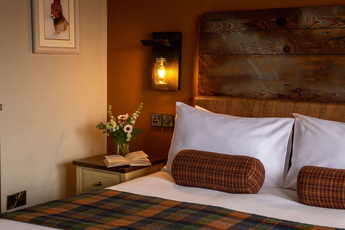 A neatly made bed with white linens and plaid pillows, a bedside table with an open book and a vase of flowers, and a rustic wooden headboard.