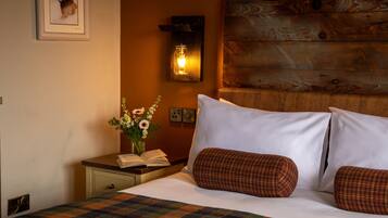 A neatly made bed with white linens and plaid pillows, a bedside table with an open book and a vase of flowers, and a rustic wooden headboard.