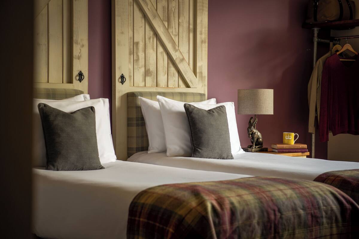 A hotel room with two beds, a wooden headboard, a small table with a lamp, and a coat rack.