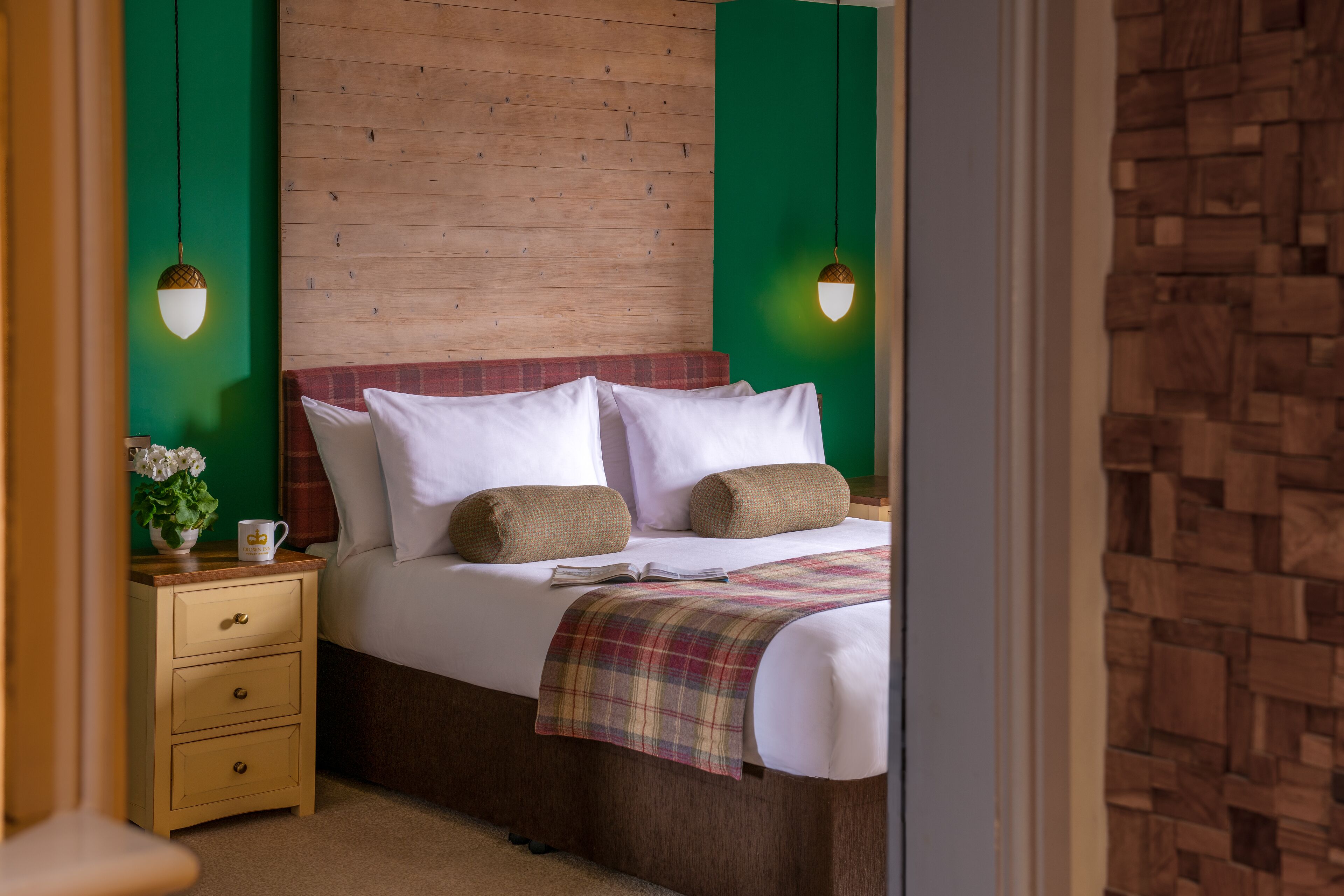 Ein ordentlich gemachtes Bett mit weißen Leinen, einer Karo-Decke und zwei Kissen. Ein Nachttisch aus Holz mit einer weißen Tasse und einer kleinen Pflanze steht daneben.