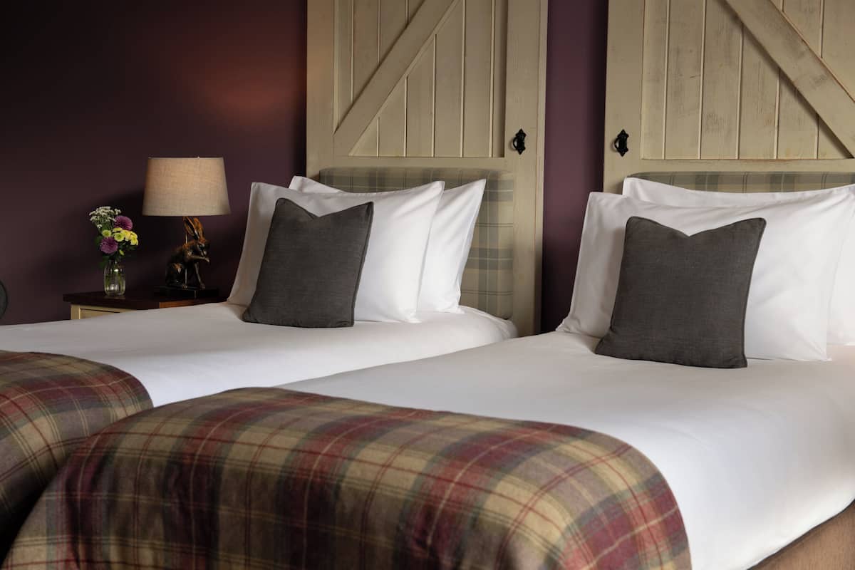 A hotel room with two beds, a lamp, a vase with flowers, and a plaid blanket.